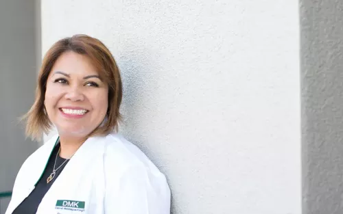 A woman with short hair and a big smile leans against a wall.