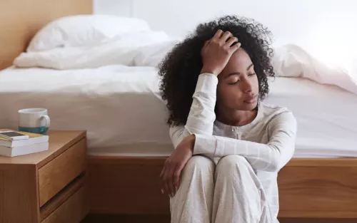A Black woman seated near a bed looking tired, holding her head with her eyes closed.