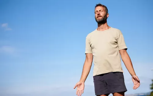 A man in running shorts and a shirt with his arms open, eyes clothed, breathing deeply.