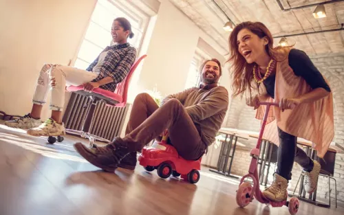 Three adults playing on some children's toy cars and scooters.