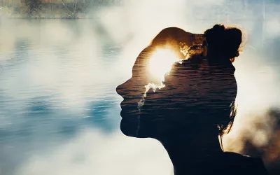 Profile outline of a person gazing outward to a body of water and a sun behind some clouds.