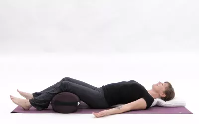 Woman on her back practicing Restorative savasana with a bolster under the knees and blanket at her head.