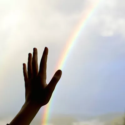 A person's hand held up to a rainbow in the sky.