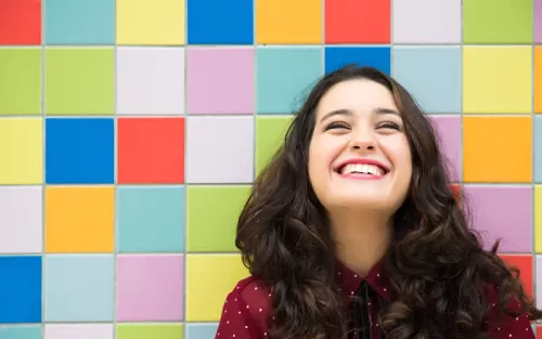 A woman with long wavy dark hair smiles in front of a wall of colorful tiles.