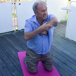 The author kneeling on a yoga mat, stretching his left elbow upward to test for flexibility.