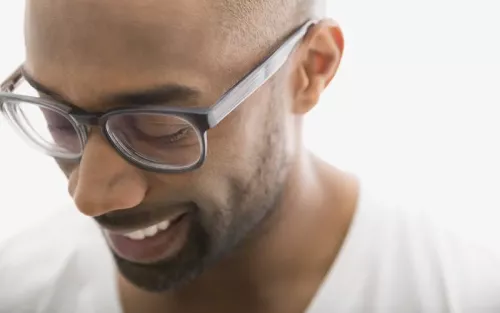 An African American man wearing clear glasses smiling and glacing downward.