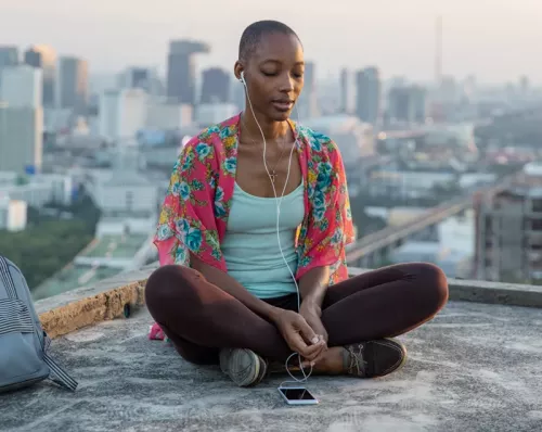A woman with a shaved head peacefully listening to music through an iPod while sitting on a rooftop.