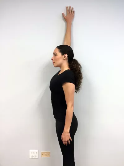 A woman with her palm of her hand above her head, pressed flat against a wall to her right side at 12 o'clock.