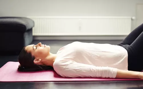 A woman lying on her back on a yoga mat in a loose white top.