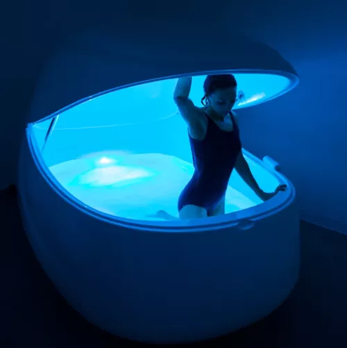 A woman in a swimsuit opens the door and climbs out of a flotation tank.