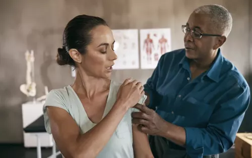 A woman pointing to her shoulder as she talks to a practitioner by her side.