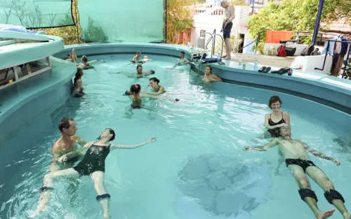 A group of 10 or 12 people doing tandem watsu in a peanut-shaped pool.