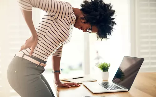African American woman with curly hair bent over a desk, holding her back in pain.