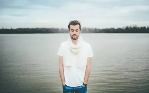 A man in a white shirt and scarf stands outside near a lake with his eyes closed.