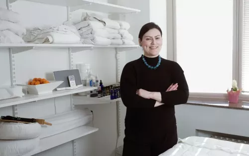 A woman in all black clothes standing in a bright white massage room.