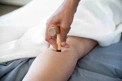 an MT's hand using a small wooden T-bar massage tool on a client's leg.