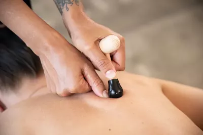 close up of two hands of a therapist using a T-bar massasge tool on a client's back with their thumbs to stabilize the tool.