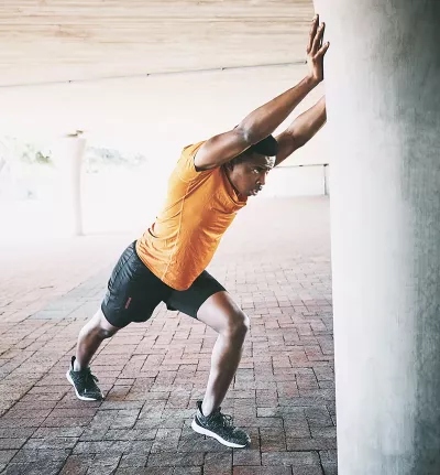 A Black man in exercise clothes pushes against a concrete column to stretch.