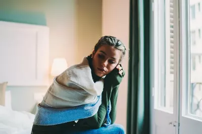 A woman sits on a bed near a window, lost in thought.