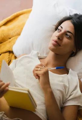 A woman lying on a pillow and blanket writes in a journal. 