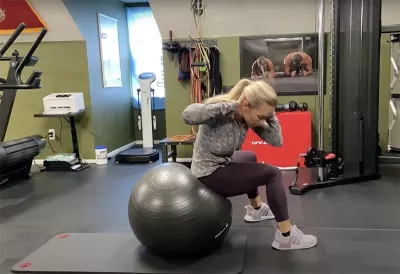 a woman demonstrating oblique crunches by sitting with her butt and back flat on the ball, twisting in a crunch.