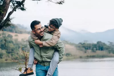 an older child rides piggyback on his father by a lake outdoors.