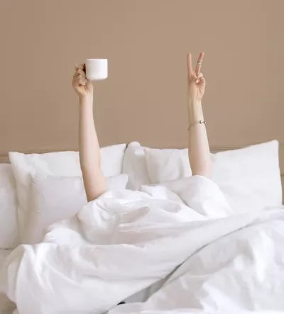 A person under the covers in bed holds up a peace sign with one hand and a coffee cup in the other hand.