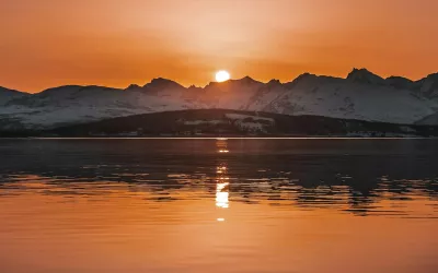 The sun sets over snowy peaks and a lake.
