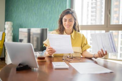A woman smiles while looking at paperwork. A calculator and tablet are on the table in front of her.