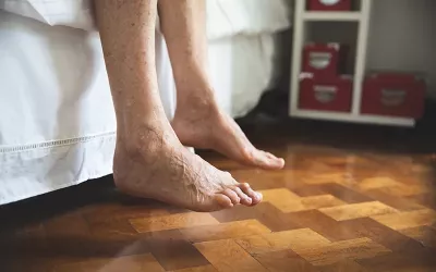 A pair of geriatric feet hover at the side of the bed and above a wooden floor.